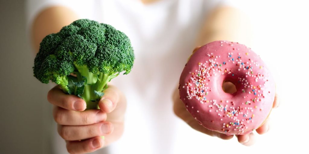 person håller i en broccoli och en munk för att illustrera hälsosamma matval